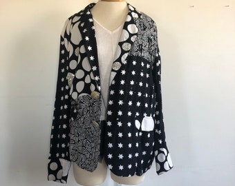 May include: A black and white patterned blazer with a white collar and a star pattern on the front. The blazer has a button closure and a pocket on the left side. The blazer is decorated with white polka dots and geometric patterns.
