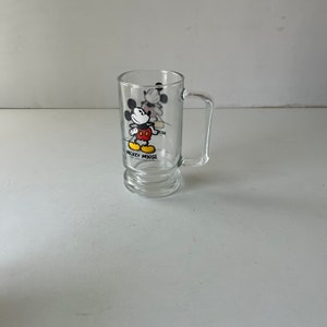 May include: A clear glass mug with a Mickey Mouse design. The mug has a handle and a rounded bottom. The design features Mickey Mouse in his signature red shorts and yellow shoes. The text "Mickey Mouse" is printed below the image.