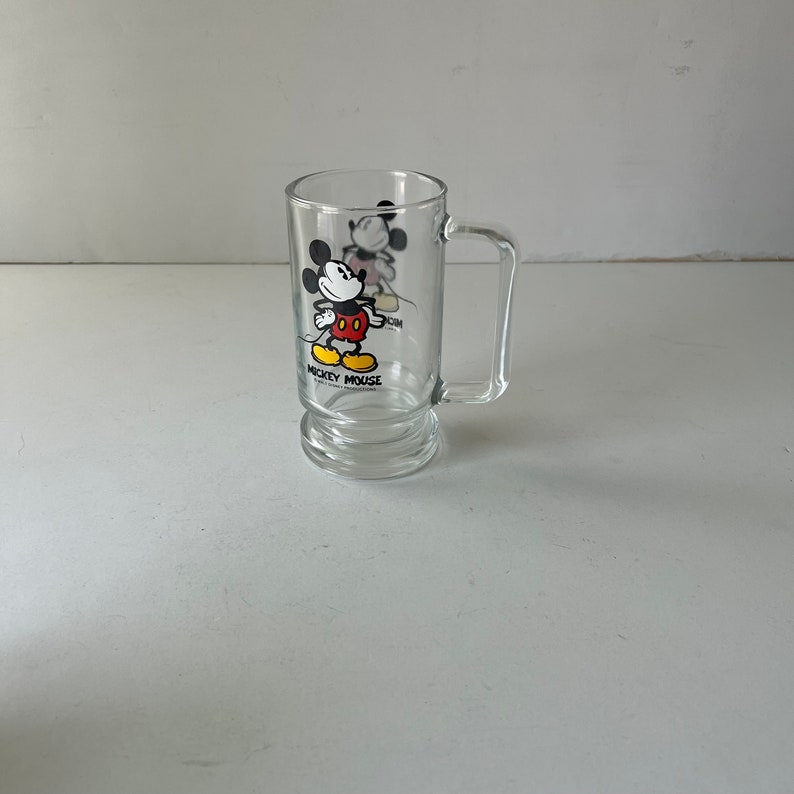 May include: A clear glass mug with a Mickey Mouse design. The mug has a handle and a rounded bottom. The design features Mickey Mouse in his signature red shorts and yellow shoes. The text "Mickey Mouse" is printed below the image.