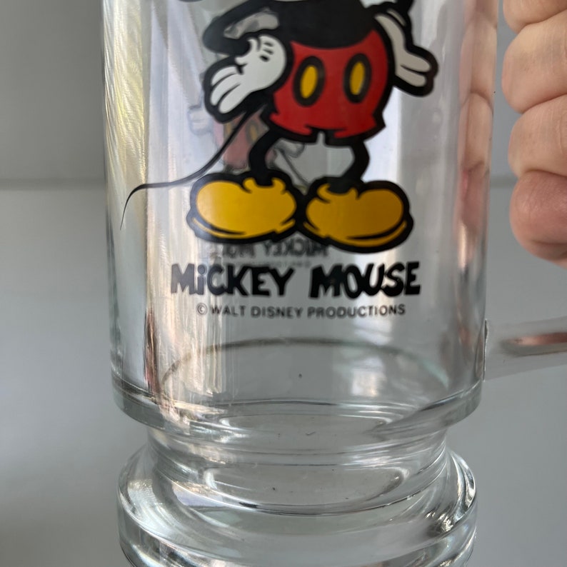 May include: A clear glass Mickey Mouse mug with a black outline and yellow shoes. The mug has a black handle and the text "Mickey Mouse" and "&copy; WALT DISNEY PRODUCTIONS" printed on the front.
