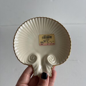 May include: A cream-colored ceramic dish shaped like a scallop shell with a gold-trimmed edge. The dish features a central sticker with a barcode and text including 118886, AEGEAN DISH, SUG. RET. 41.00, SAVE 32%, and NOW 27.95 printed on the surface.