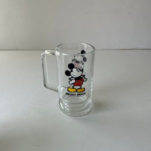 May include: A clear glass mug with a handle features a cartoon image of Mickey Mouse in his signature red shorts and yellow shoes. The mug has the text "Mickey Mouse" printed on the bottom.