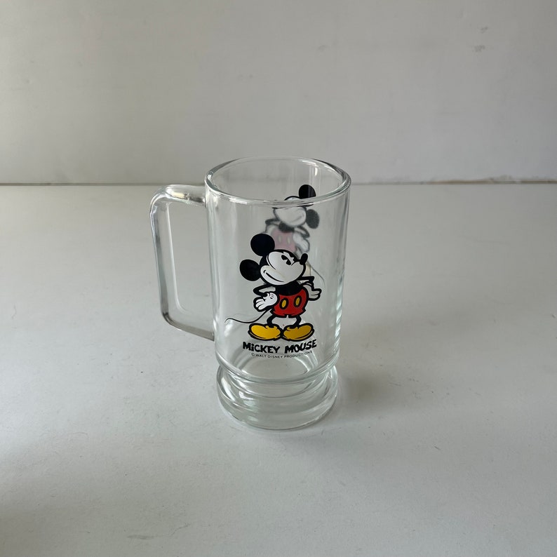 May include: A clear glass mug with a handle features a cartoon image of Mickey Mouse in his signature red shorts and yellow shoes. The mug has the text "Mickey Mouse" printed on the bottom.