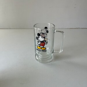 May include: A clear glass mug with a Mickey Mouse design. The mug features a black and white illustration of Mickey Mouse wearing red shorts and yellow shoes. The text "Mickey Mouse" is printed below the illustration.