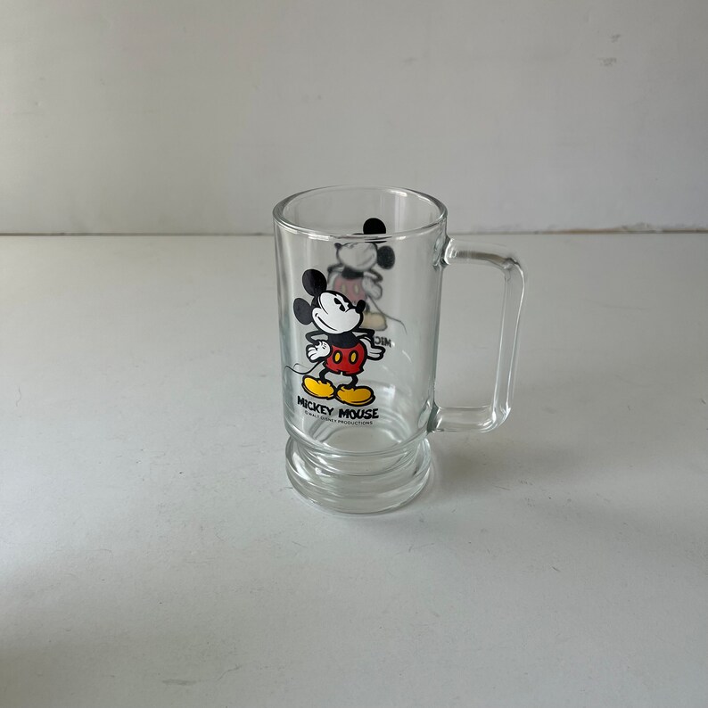 May include: A clear glass mug with a Mickey Mouse design. The mug features a black and white illustration of Mickey Mouse wearing red shorts and yellow shoes. The text "Mickey Mouse" is printed below the illustration.