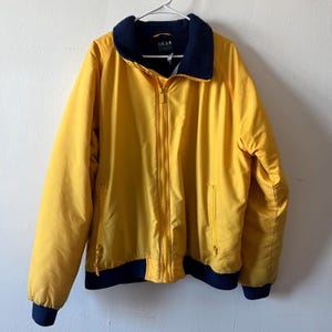 May include: A yellow jacket with a navy blue lining and zipper. The jacket has a logo that says "GEAR" on the front.