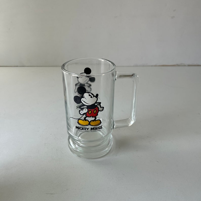 May include: A clear glass mug with a black and white image of Mickey Mouse. The mug has a handle and a rounded base. The text "Mickey Mouse" is printed on the mug.