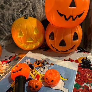 May include: A collection of orange plastic jack-o'-lanterns including two large buckets, one round pumpkin, a small pumpkin-topped flashlight, and two miniature pumpkin charms resting on a light blue plastic bag with ghost illustrations.