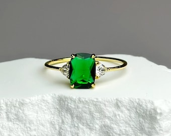 Op de afbeelding: Een gouden ring met een grote smaragdgroene edelsteen en twee kleinere heldere edelstenen. De ring ligt op een witte steen.