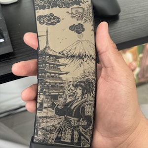 May include: A black and tan patterned magazine with a Japanese-inspired design. The design features a mountain, a pagoda, clouds, cherry blossoms, and a woman holding a mask.