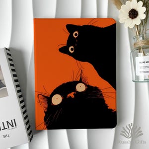 May include: An orange tablet case with a cartoon illustration of two black cats with large, surprised eyes. The cats are set against a bright orange background. The case is a protective accessory for tablets.