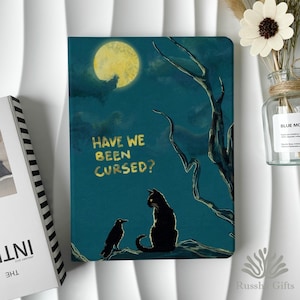 May include: A notebook with a dark teal cover featuring a black cat and raven under a full moon. The text "HAVE WE BEEN CURSED?" is in gold lettering. A whimsical, spooky design.