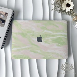 Op de afbeelding: Een laptop met een groen en roze marmerpatroon hoes, met het Apple-logo. De laptop staat op een wit oppervlak, met een boek en een klein boeket bloemen. De hoes heeft een glanzende afwerking.