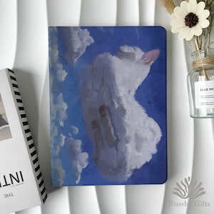 May include: A notebook with a blue cover featuring a white cat illustration resembling a cloud against a blue sky. The notebook is next to a book with the word "INT" visible and a vase with a flower.