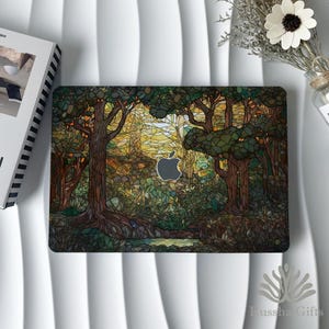 May include: A laptop skin featuring a forest scene with trees, foliage, and a yellow sky. The design has a stained-glass effect, with the Apple logo visible. The skin is on a white surface, with a book and flowers nearby.