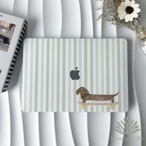 May include: A laptop skin featuring a light sage green and white vertical striped pattern. A brown dachshund dog is illustrated riding a pink skateboard with yellow wheels positioned in the lower right section of the rectangular surface.