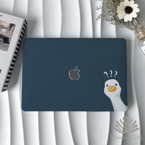 May include: A navy blue laptop case featuring the Apple logo and a cartoon goose with question marks. The goose has a white head, yellow beak, and is peeking from the side. The case is on a white surface.