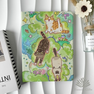 Puede incluir: Una funda para tableta con una ilustración caprichosa de tres gatos de dibujos animados sobre un fondo verde y azul con detalles florales y de estrellas. Los gatos están en varias poses, incluido un gato naranja y blanco sentado.