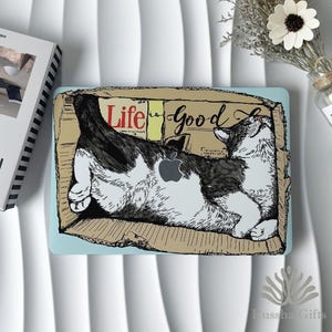 May include: A laptop case featuring a black and white cat design inside a cardboard box with the words "Life is Good". The Apple logo is visible on the cat's chest. The case has a light blue border.