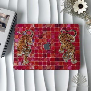 May include: A rectangular laptop skin featuring a grid of red and orange square tiles. Two illustrated tigers with orange and black stripes are positioned on the left and right sides, facing inward toward a central silver Apple logo cutout.