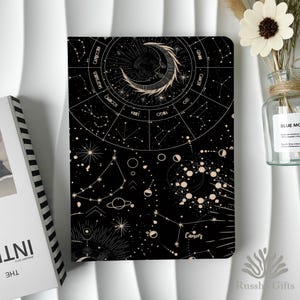 May include: Black notebook featuring a celestial design with constellations, zodiac symbols, and a crescent moon. The notebook has a detailed astrological chart and the word "Cancer" is visible. The notebook is next to a vase with a white flower.