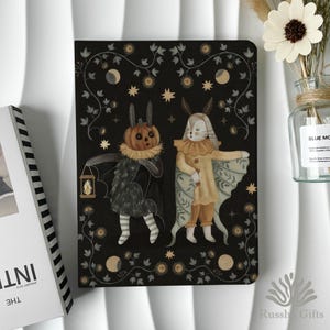 May include: A black notebook featuring a whimsical illustration of two costumed figures, one with a pumpkin head and the other with rabbit ears. The design includes stars, moons, and floral elements. The notebook is next to a vase with a white flower.
