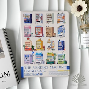 May include: A white notebook featuring a colorful illustration of various vending machines. The cover includes the text "THE VENDING MACHINE CATALOGUE." A vase with a white flower and a book are in the background.