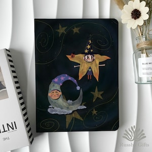 Pode incluir: Um caderno com uma ilustração fantasiosa de uma figura em forma de estrela usando um chapéu de bruxa, uma lua crescente com um rosto adormecido e estrelas em um fundo escuro. O caderno tem um design em espiral e o texto "Russ Gifts" está na parte inferior.