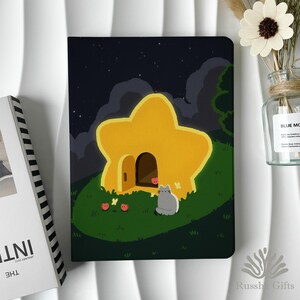 May include: A rectangular notebook featuring a dark night sky illustration with a large, yellow, star-shaped house on a green grassy hill. A small gray cat sits in front of the open door, surrounded by tiny red apples and yellow butterflies.
