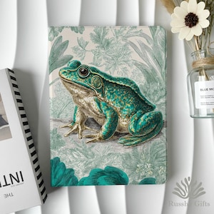 Op de afbeelding: Een notitieboek met een gedetailleerde illustratie van een groene en gouden kikker. De kikker staat tegen een achtergrond van groen gebladerte. De kaft van het notitieboek heeft een teal en beige kleurenschema, met de tekst "Januari 2022" zichtbaar op de rug van een boek.