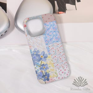 Op de afbeelding: Een telefoonhoesje met bloemenpatroon en patchwork-ontwerp. De hoes heeft verschillende bloempatronen in blauw, roze en wit. Het heeft uitsparingen voor de camera en knoppen. De merknaam "Russha Gifts" is zichtbaar.