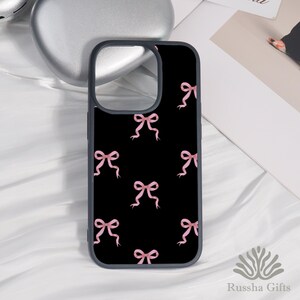 Op de afbeelding: Zwarte telefoonhoes met een patroon van roze aquarel strikken. De hoes heeft een donkergrijze rand en een uitsparing voor de camera. De telefoonhoes ligt op een witte ondergrond.