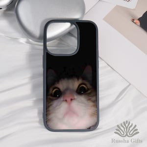 May include: A black phone case with a close-up image of a cat's face. The cat has large, wide eyes and a pink nose. The case has a dark background and a gray border. The brand name "Russha Gifts" is at the bottom.