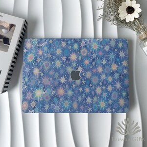 May include: A laptop with a blue cover featuring a pattern of colorful starbursts. The Apple logo is visible on the cover. The laptop is on a white surface, with a book and flowers nearby.