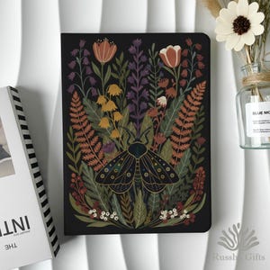 May include: A black journal with a colourful floral and moth design. The cover features a detailed illustration of a moth surrounded by various flowers and foliage in shades of orange, green, and purple. The journal is next to a vase with flowers.
