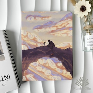 May include: A fantasy-themed journal with a vibrant illustration of a dragon and a person on a mountain against a sunset sky. The cover art features a color palette of purple, orange, and yellow. The journal is rectangular and appears to be made of a smooth material.
