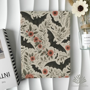 May include: A beige notebook with a repeating pattern of black bats, red flowers, and green leaves. The notebook is next to a vase with a white flower and a book with the word "INT" visible.