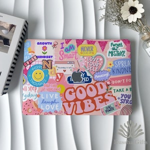 May include: A pink laptop case with a collage of colorful stickers. The stickers feature phrases like "Good Vibes," "Love Yourself," and "Never Give Up." Other stickers include a smiley face, an apple, and a donut.