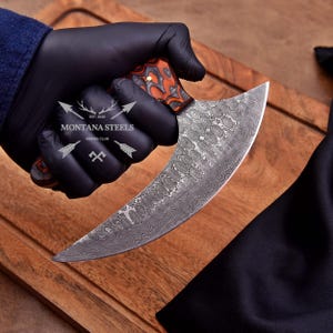May include: A curved, Damascus steel knife with a patterned blade and a handle featuring orange and brown accents. The knife is held in a gloved hand, with the "MONTANA STEELS KNIVES CLUB" logo visible. The knife has a curved blade.
