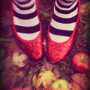 May include: A pair of red glitter shoes with black and white striped socks. The shoes are in a field of green grass with fallen leaves and red apples.