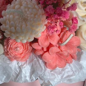 May include: A close-up of a floral arrangement featuring various flower-shaped candles in shades of pink and white. The arrangement includes a large, cream-colored chrysanthemum candle, pink rose candles, and smaller pink flower candles, all nestled in a pink container.