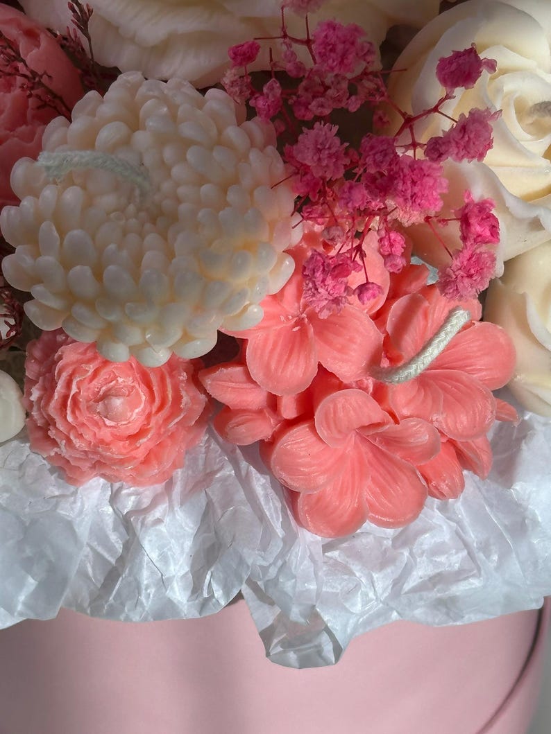 May include: A close-up of a floral arrangement featuring various flower-shaped candles in shades of pink and white. The arrangement includes a large, cream-colored chrysanthemum candle, pink rose candles, and smaller pink flower candles, all nestled in a pink container.