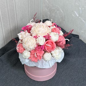 May include: A floral arrangement of flower-shaped candles in a pink hatbox. The candles are in shades of white, pink, and coral, with small pink flowers and sprigs of dried pink flowers. The hatbox is round and pink, with white tissue paper.