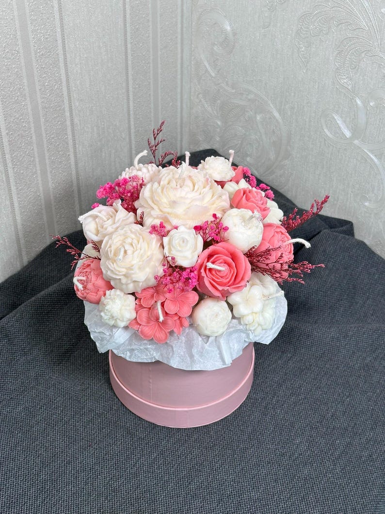 May include: A floral arrangement of flower-shaped candles in a pink hatbox. The candles are in shades of white, pink, and coral, with small pink flowers and sprigs of dried pink flowers. The hatbox is round and pink, with white tissue paper.