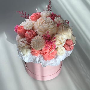 May include: A floral arrangement of carved candles in a pink round box. The candles are shaped like various flowers in shades of white, pink, and coral. The arrangement includes roses, peonies, and other blossoms, with small pink accents.