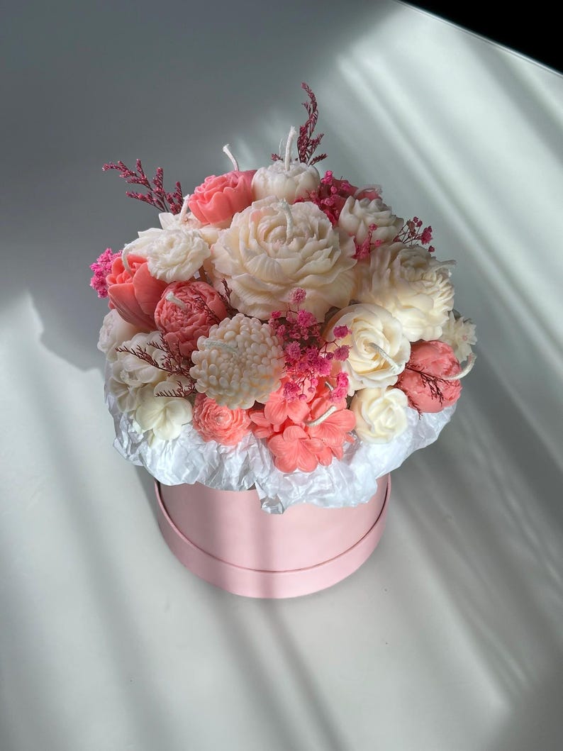 May include: A floral arrangement of carved candles in a pink round box. The candles are shaped like various flowers in shades of white, pink, and coral. The arrangement includes roses, peonies, and other blossoms, with small pink accents.