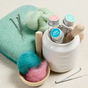 May include: A white ceramic jar with three glass vials containing felting needles. The vials are labeled "38S Medium", "10T Fine", and "36T Coarse".  There are also two wooden spoons with pink and teal wool roving, and three felting needles on a green felting pad.