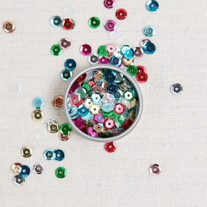 May include: A silver container filled with colorful sequins in shades of blue, green, red, and gold. The sequins are scattered around the container on a white fabric background.