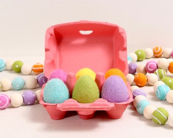 May include: A pink cardboard egg carton filled with six colorful felt Easter eggs. The eggs are in shades of green, blue, pink, and purple. The carton is sitting on a white surface.