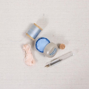 May include: An assortment of sewing supplies, including a spool of light blue thread, a bundle of pink thread, a blue wax container, a cork, and a needle tool. The items are arranged on a light-colored fabric background.